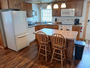 A kitchen with wooden cabinets, a refrigerator, a microwave, a stove, and a sink. There are two wooden chairs at the island.