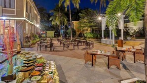 An outdoor dining area with tables and chairs, surrounded by palm trees and a building with a balcony.