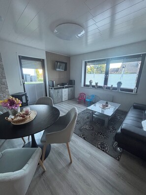 Eine moderne Küche mit Einbauofen, eine Essecke mit rundem Tisch und Stühlen, eine Couch und ein Fenster mit Jalousien.