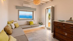 Una habitación con un ventanal grande con vistas al mar, un sofá con cojines, un aparador de madera y una lámpara colgante.