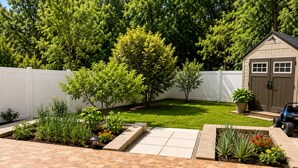 Ein Garten mit Terrasse, Gartenhaus und weißem Zaun.