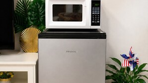 Ein Hisense-Kühlschrank mit einer Mikrowelle darüber, einem Fernseher links und einer Topfpflanze rechts.