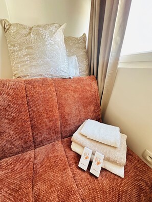 Ein Zimmer mit einem roten Sofa, zwei Kissen, einem Fenster mit Vorhängen und einem Badeköffchen auf dem Sofa.