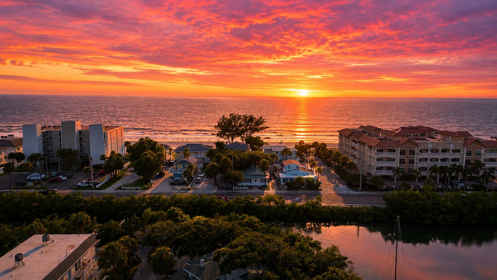 A coastal town at sunset with buildings, a marina, and a clear view of the ocean.