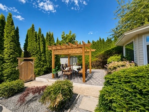 A wooden pergola with a table and chairs, surrounded by well-maintained hedges and trees.