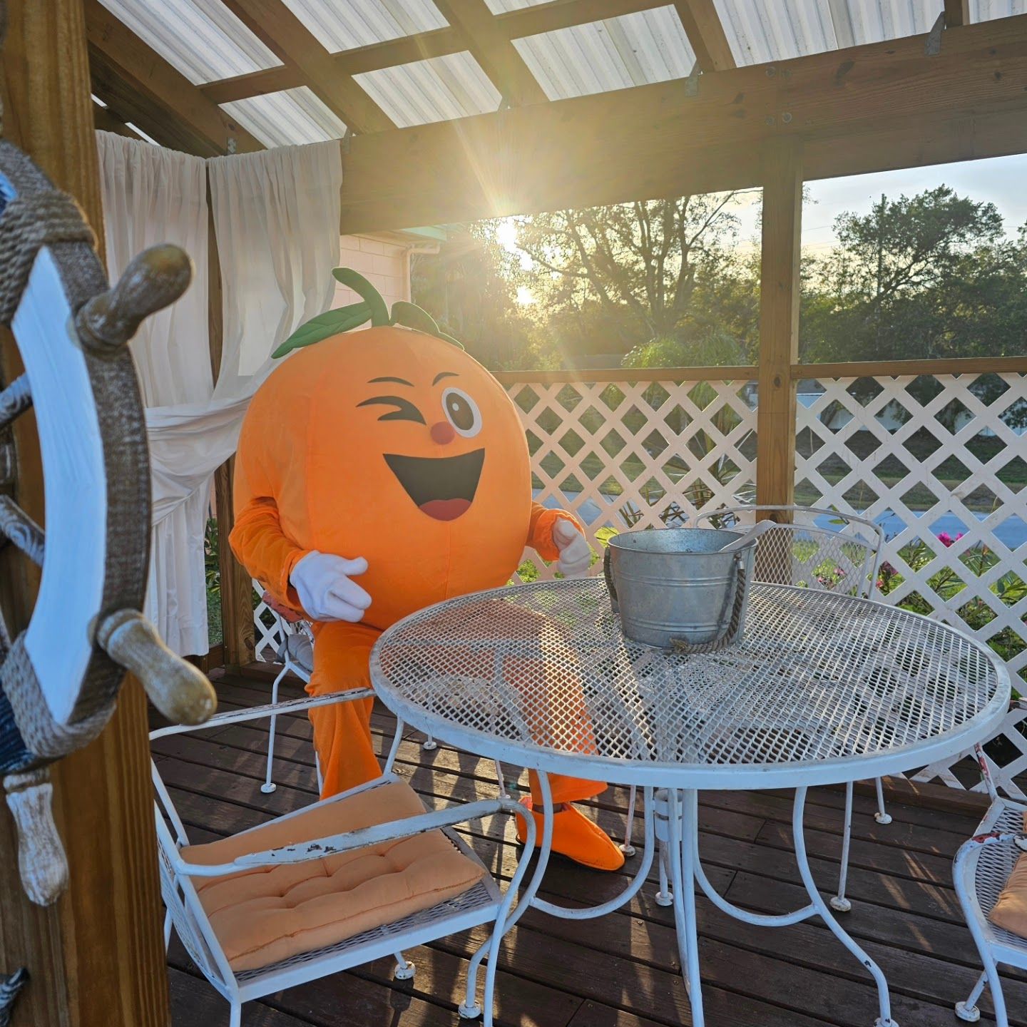 A person in a pumpkin costume sitting at a round metal table with a metal bucket on it, on a wooden deck with a lattice fence in the background.