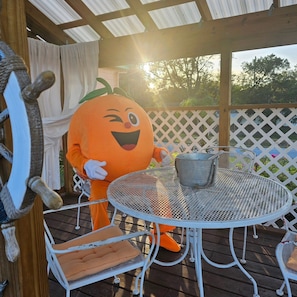 Una persona disfrazada de calabaza sentada a una mesa metálica redonda con un cubo de metal encima, en una terraza de madera con una valla de celosía al fondo.
