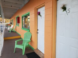 Een motel met een veranda, groene stoelen en een witte deur met een nummer.
