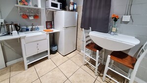 A compact kitchen with a white countertop, a microwave, a refrigerator, a sink, and a small dining area with a chair and table.