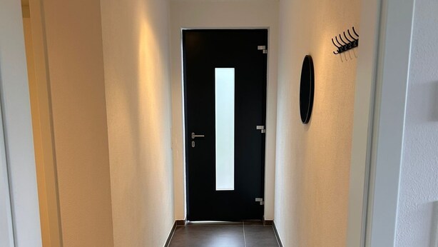 Ein Flur mit einer schwarzen Tür am Ende, einem kleinen runden Gegenstand an der Wand und einer Lüftungsgitter rechts.