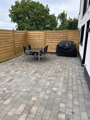 Eine Terrasse mit Tisch und Stühlen, ein überdachter Grill und ein Holzzaun.