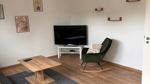 Ein Wohnzimmer mit Fernseher, einem grünen Sessel, einem Holztisch und einer Couch.