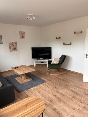 Ein modernes Wohnzimmer mit Couchtisch aus Holz, einem grünen Sessel, einem Flachbildfernseher und einer weißen Tür.