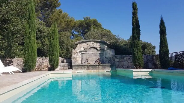 Ein Schwimmbecken mit einer Steinbrunnen, Liegestühlen und hohen Zypressen.