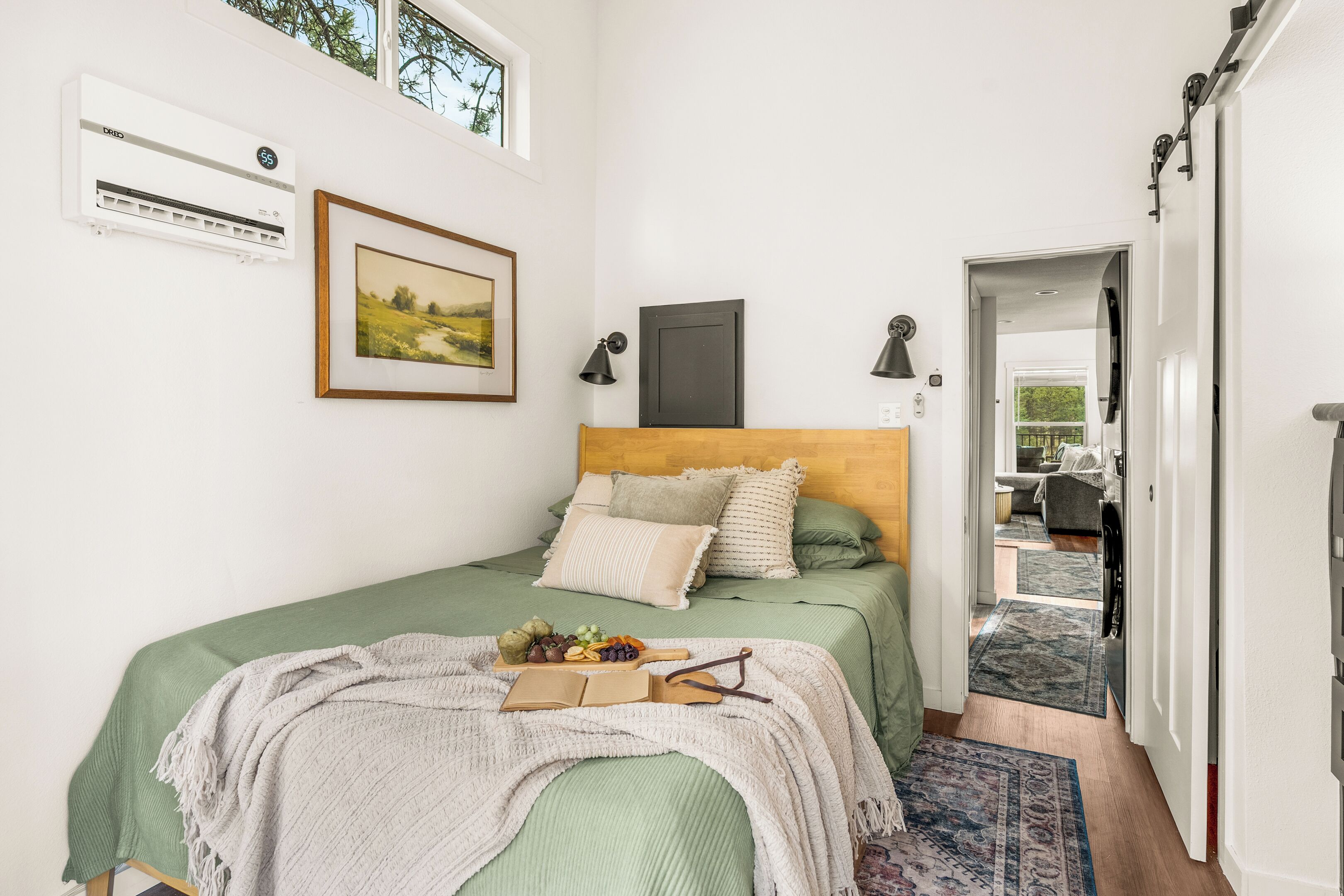 Een slaapkamer met een bed, nachtkastje met een boek en brillen, een ingelijste foto, een airconditioner en uitzicht op de aangrenzende kamer.