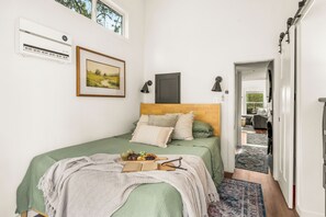 A bedroom with a bed, a bedside table with a book and glasses, a framed picture, an air conditioning unit, and a view into the adjacent room.
