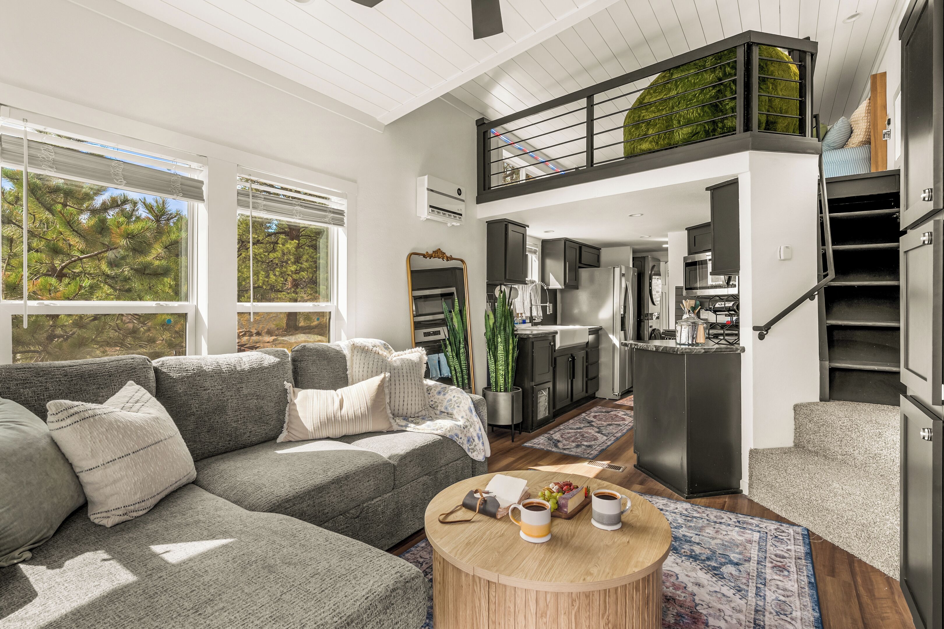 A modern living room with a gray sectional sofa, a wooden coffee table with cups, a kitchen area with dark cabinets, and a balcony with green plants.