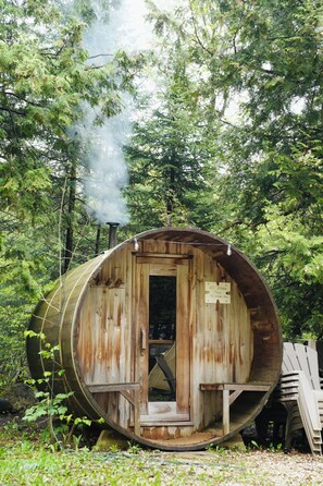 Une petite maison rustique en bois, construite à partir d’un tonneau, avec une porte, un petit porche et une cheminée d’où s’échappe de la fumée.