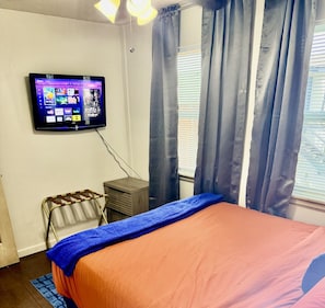 Ein Schlafzimmer mit Bett, Wandfernseher, kleinem Schreibtisch und einem Fenster mit Vorhängen.