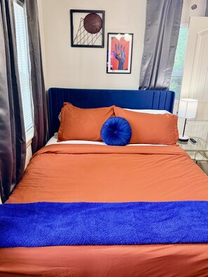 A bed with orange bedding and a blue accent pillow, a blue headboard, and two framed artworks on the wall. - Thunder Up Loft Garage Apt 10 min to Downtown (Oklahoma City)