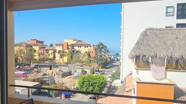 Un balcon avec une table ronde en verre et des chaises, une structure à toit de chaume et une vue sur une ville côtière.