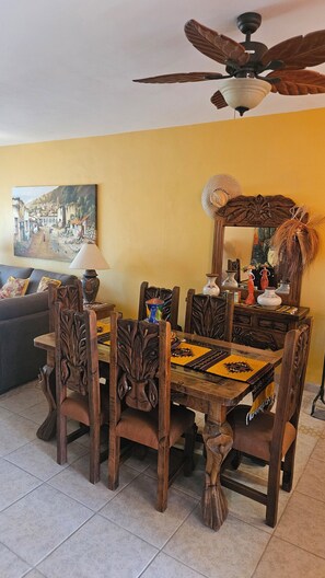 A dining area with a wooden table and chairs, a ceiling fan, a painting on the wall, and a sofa in the background. - Cozy 2-bedroom condo in perfect Bucerías with AC,  private rooftop bar area (Bucerías)