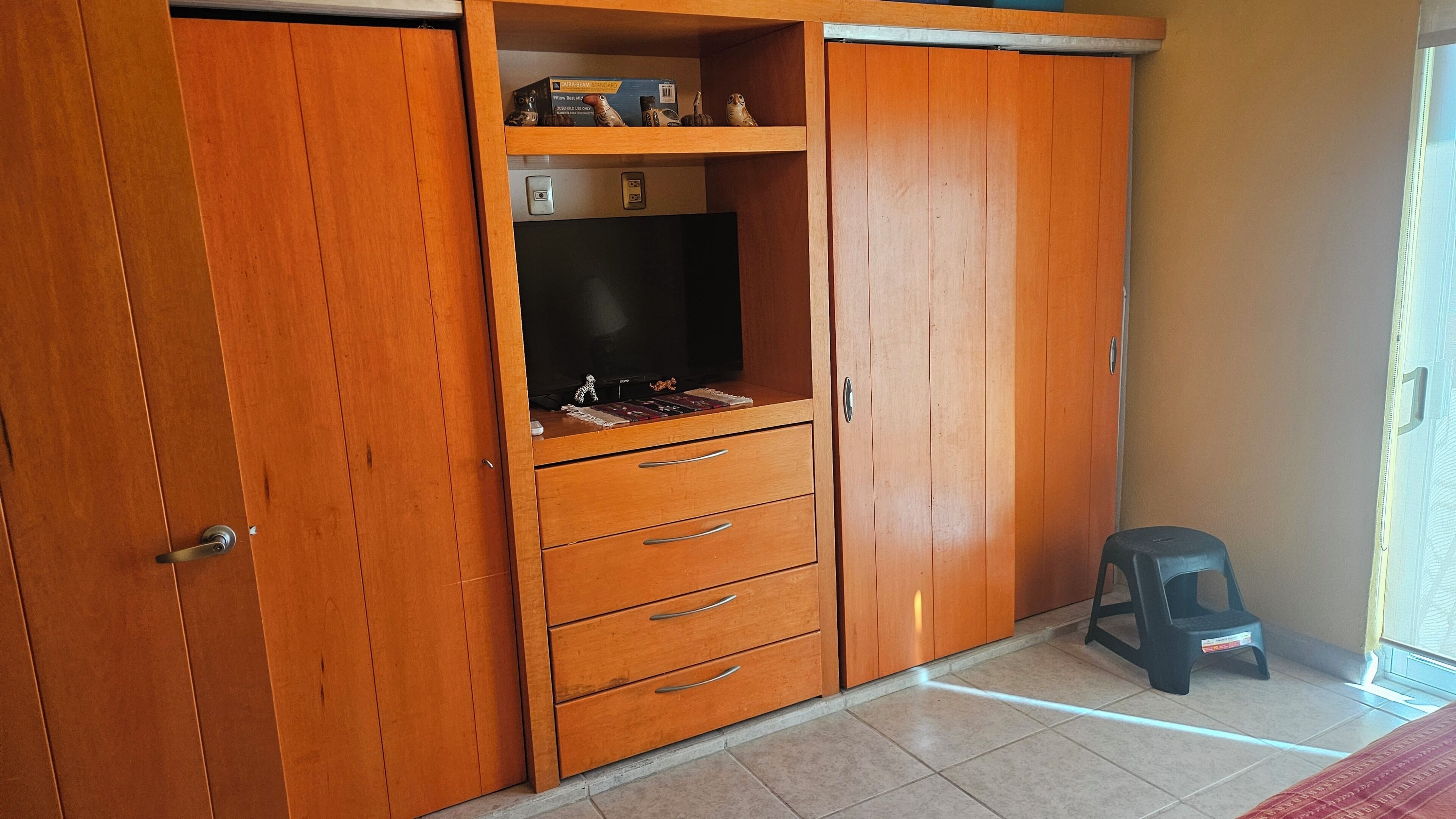 Un armario de madera con un televisor, una pequeña estantería con adornos y un taburete de plástico.