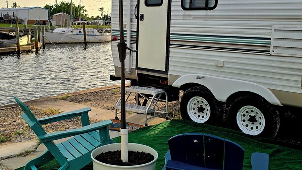 Un espace de loisirs aménagé avec une remorque, un parasol turquoise, un pot de fleurs et deux fauteuils Adirondack.