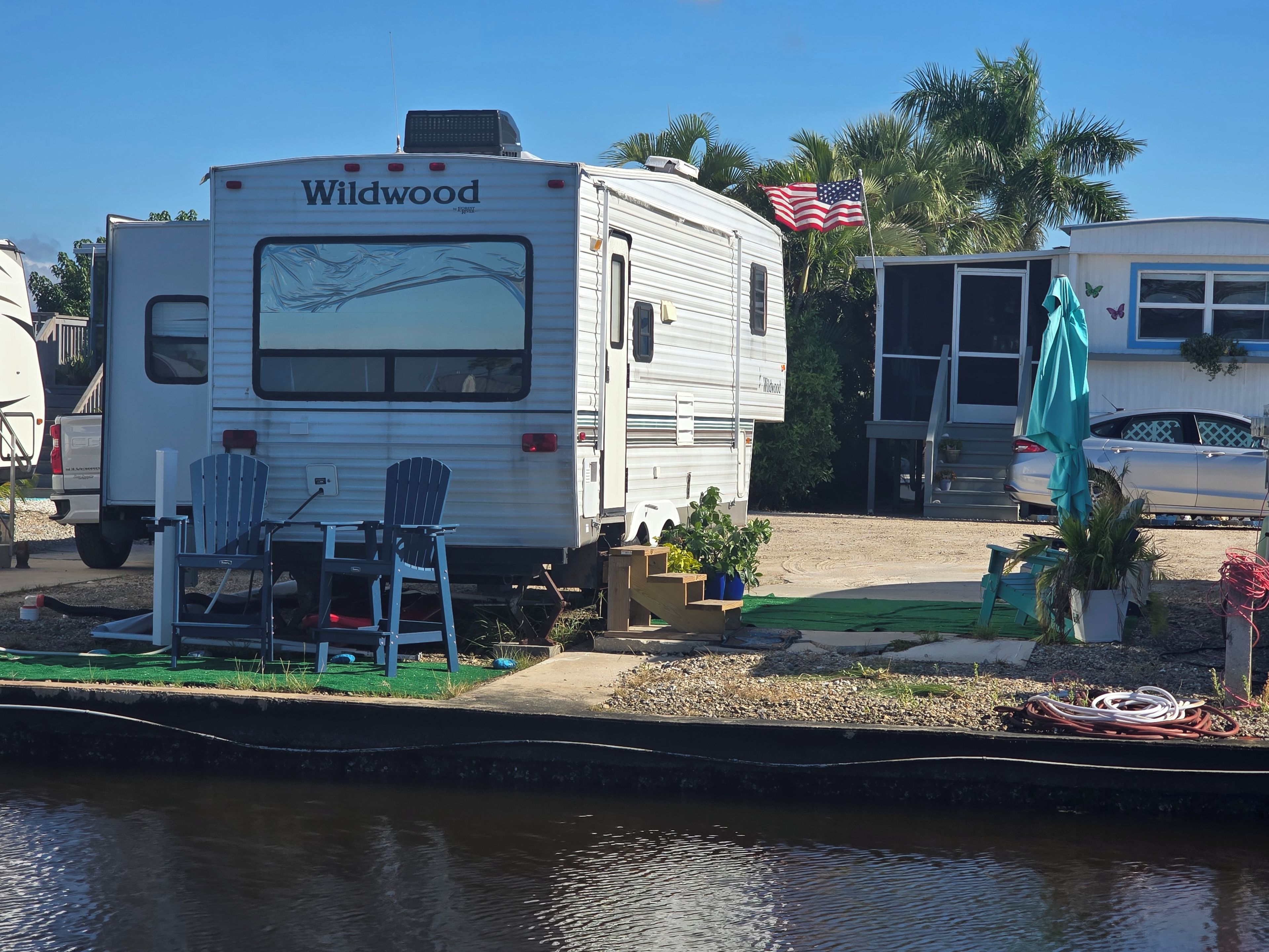 A white RV named 'Wildwood' parked near a body of water, with a small deck area and outdoor seating.