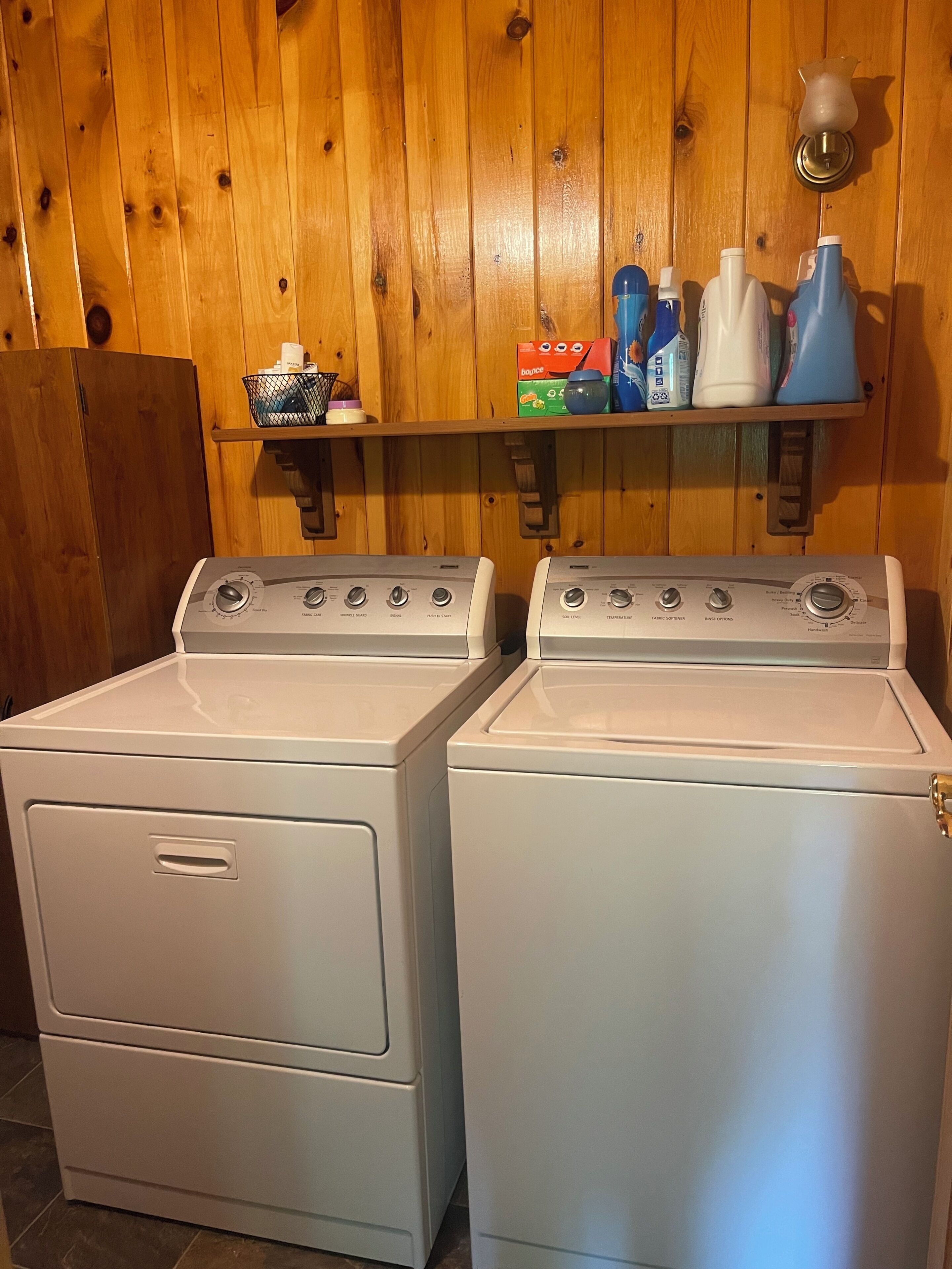 Two white front-loading washers with control panels, a wooden wall, and a shelf with cleaning supplies.