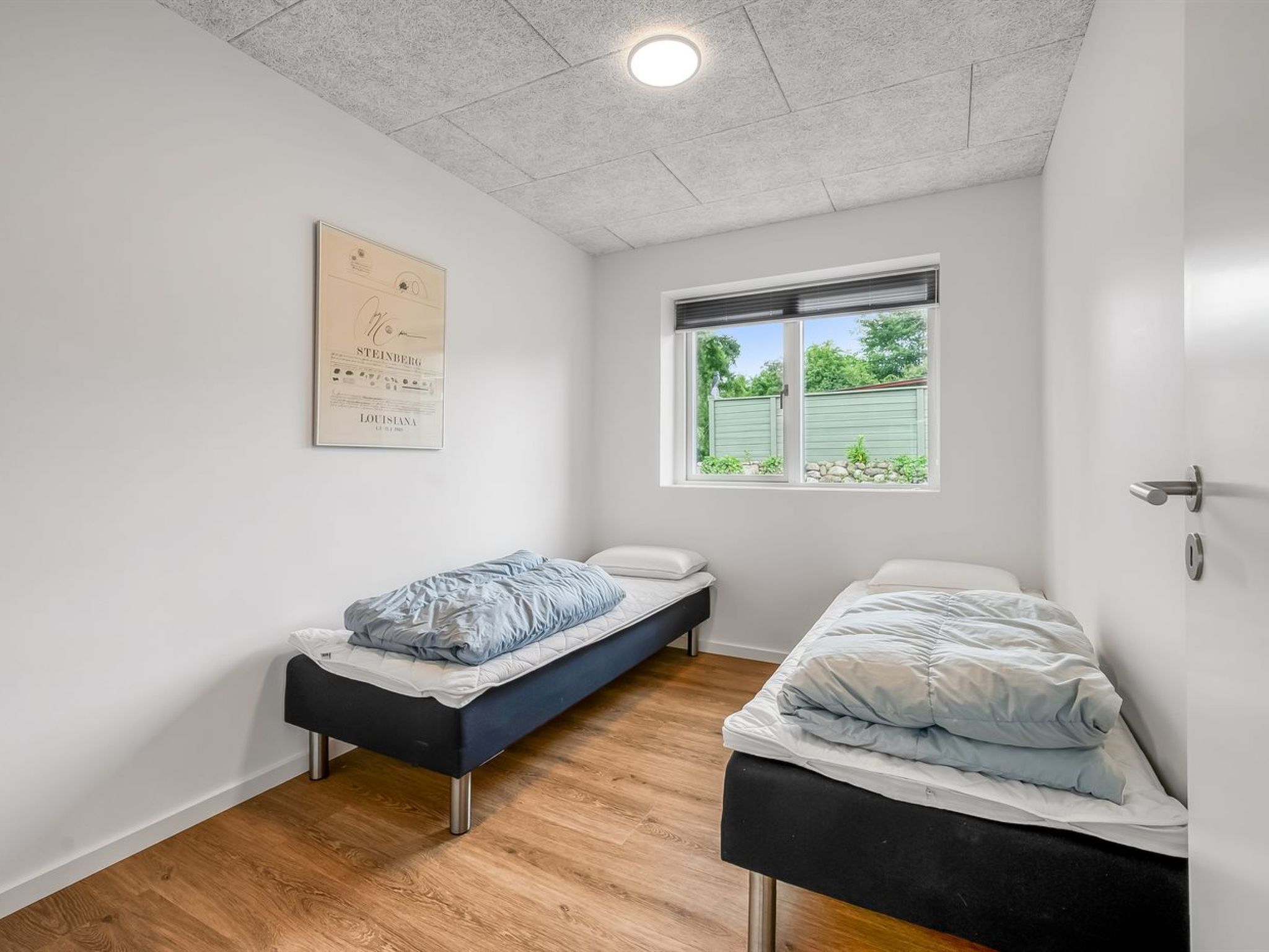 A small, clean room with two beds, a window with a view of greenery, and a ceiling light.