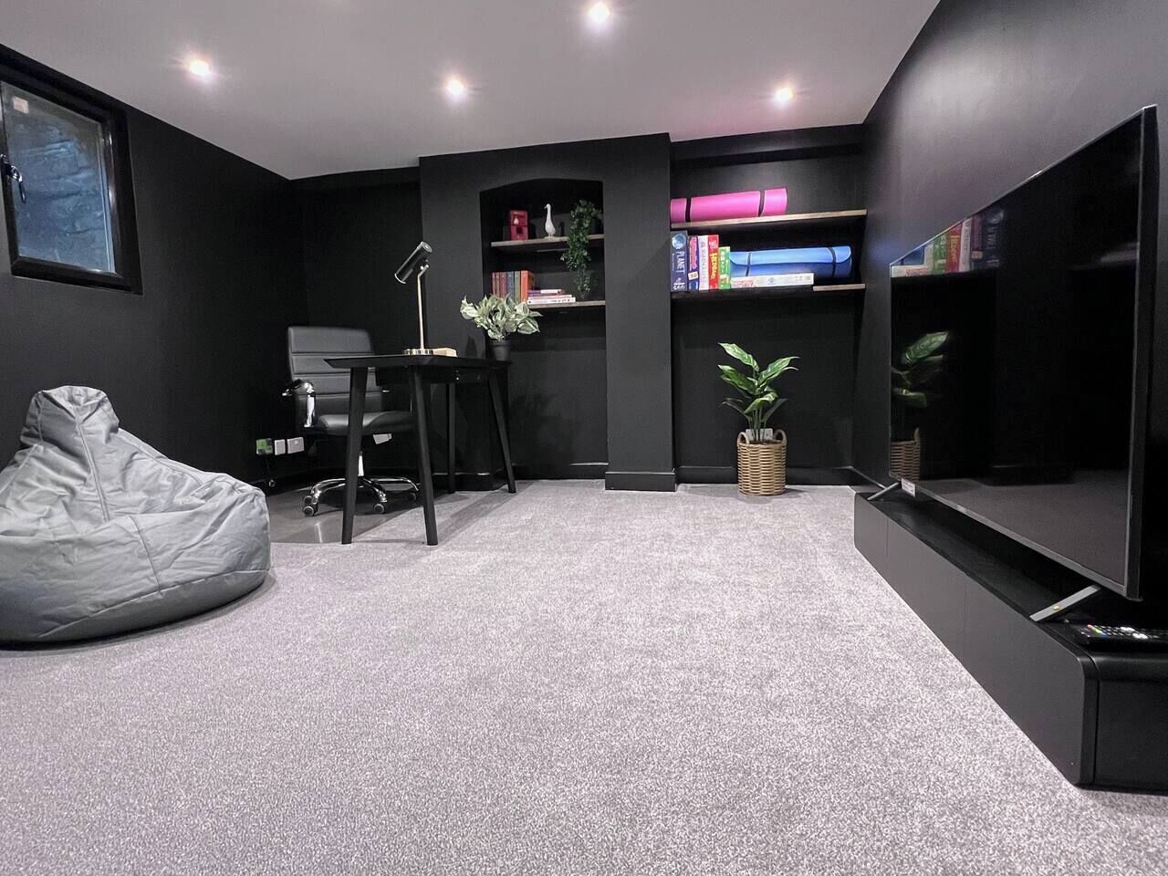 A modern living room with a grey bean bag, a desk with a lamp, a bookshelf, and a flat-screen TV.