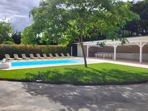 Ein Schwimmbad mit Liegestühlen, einer überdachten Terrasse und einem gepflegten Rasen.