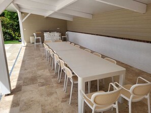 Eine überdachte Terrasse mit einem langen Tisch und Stühlen, einer kleinen Bar und Blick auf Grünflächen.