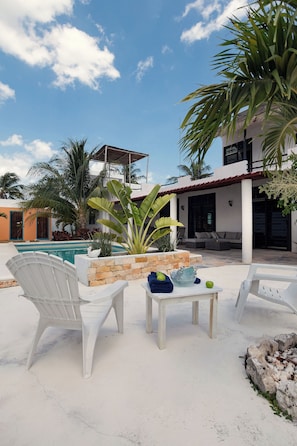 A poolside area with white lounge chairs, a small table with fruits, and a stone fire pit. - Private Pool + Beach Access + Workspace | 3BR Sleeps 6 (Chelem)