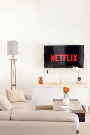 Ein modernes Wohnzimmer mit einem Fernseher, der 'NETFLIX' anzeigt, einem Sofa, einer Stehlampe, einem Couchtisch und einer Vase mit Blumen.