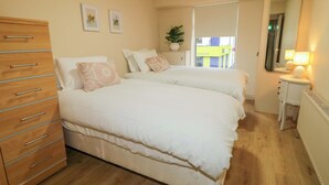 A bedroom with two beds, a wooden dresser, a bedside table, a mirror, and a potted plant. - Suite 33A (Londonderry)