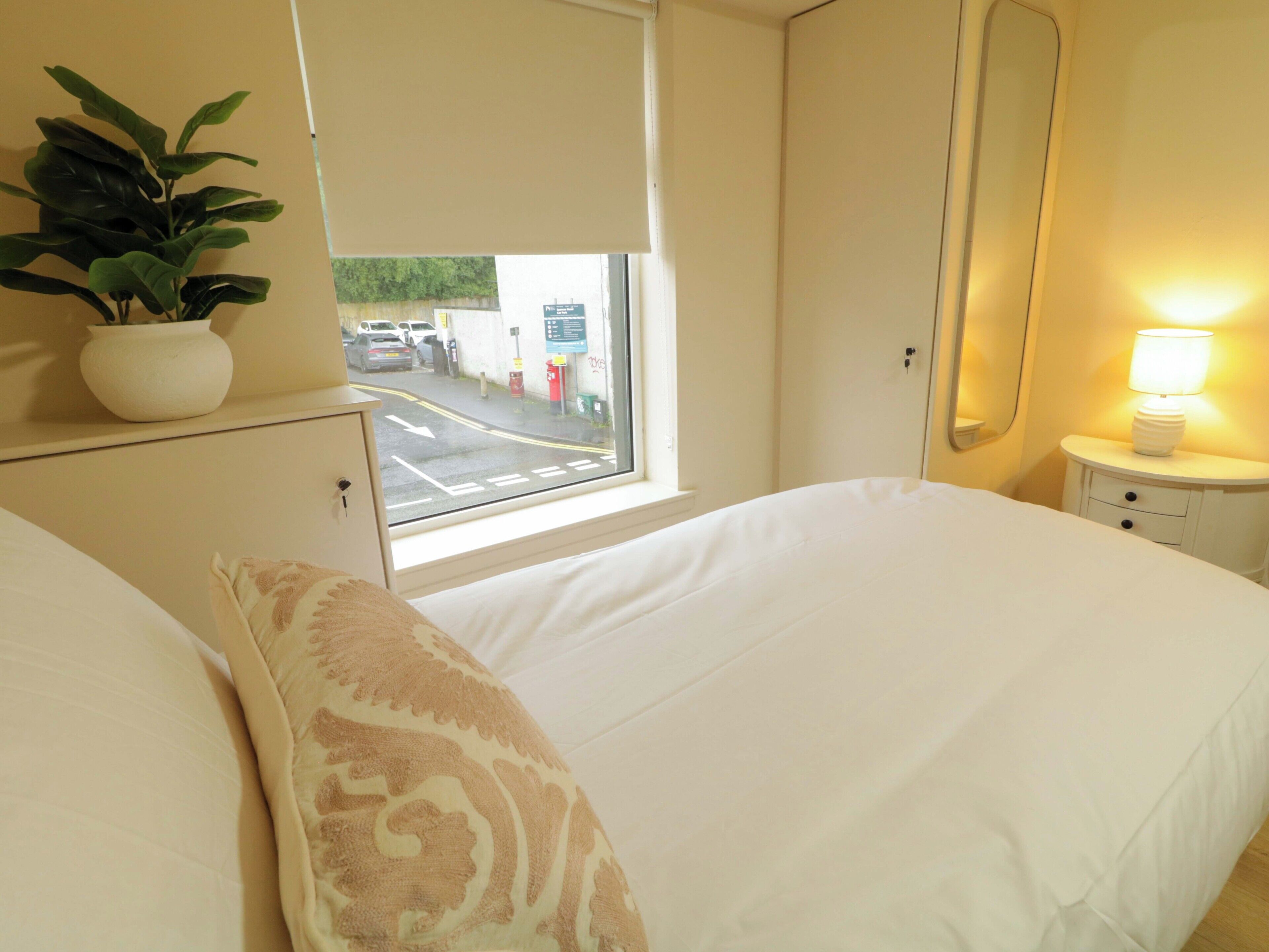 A hotel room with a bed, a bedside table with a lamp, a potted plant, and a window with a view of the street.