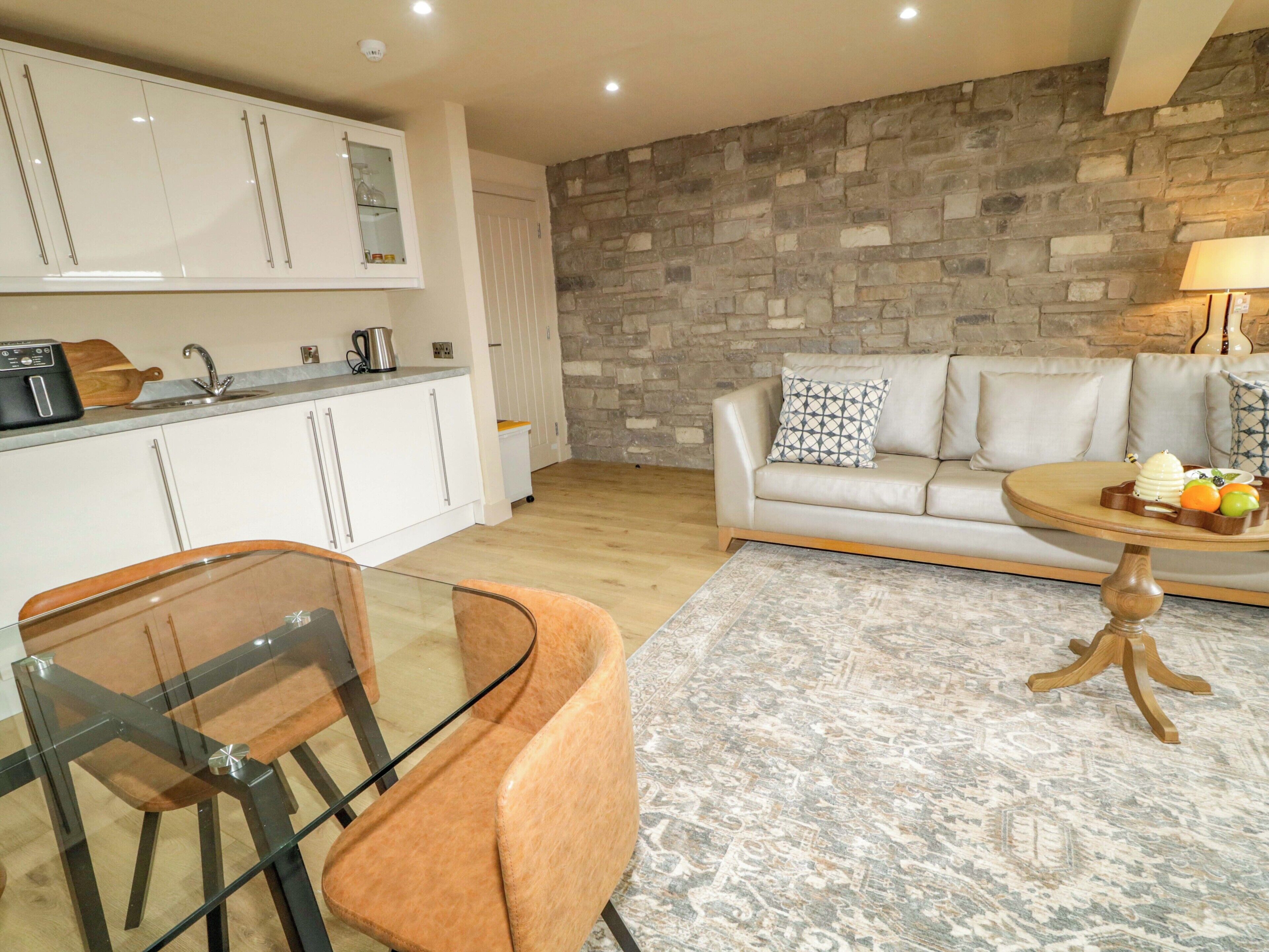 A modern kitchen with white cabinets, a stone wall, a beige sofa, and a dining area with a glass table and orange chair.