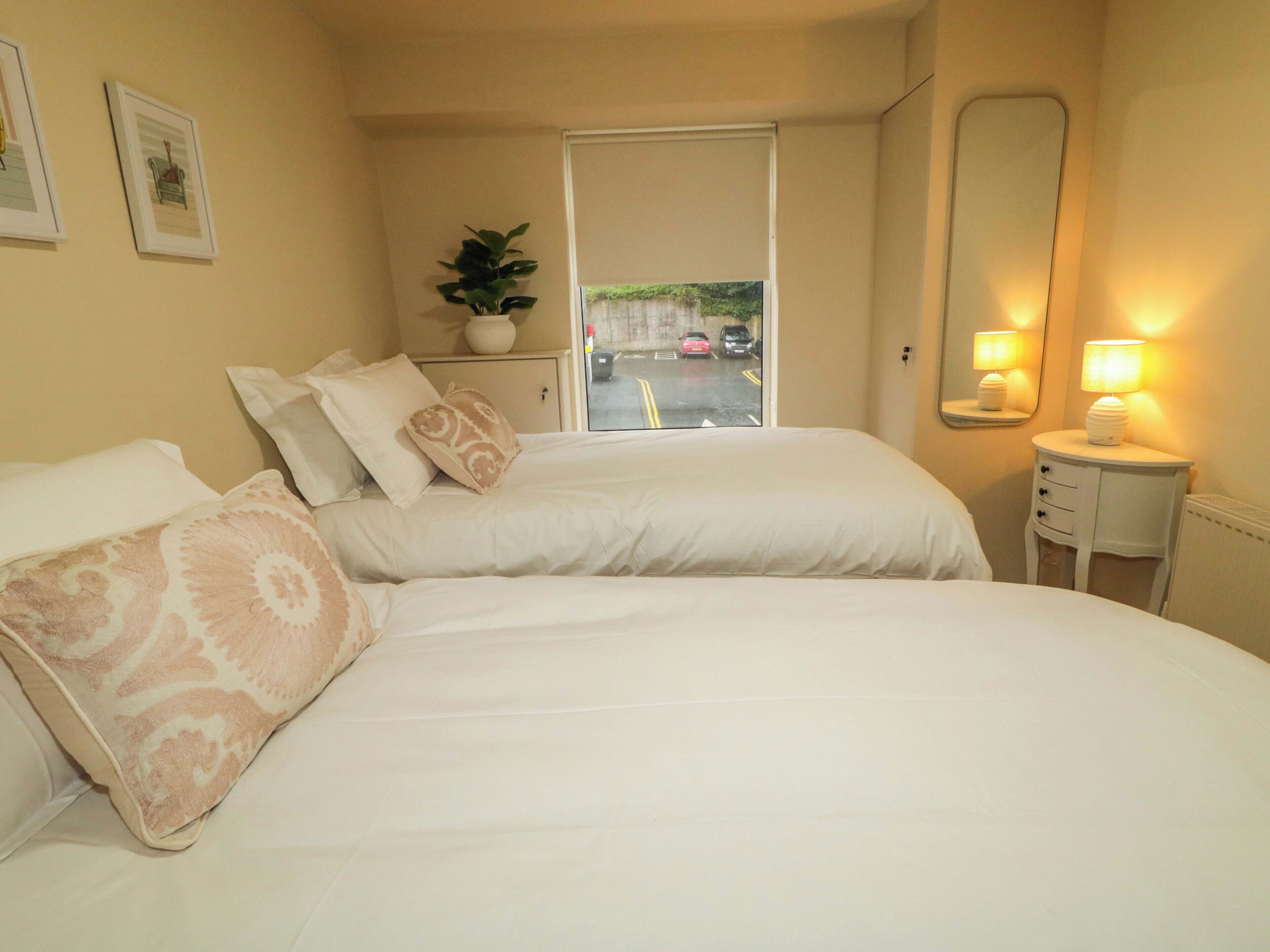 A hotel room with two beds, a potted plant, a bedside table with a lamp, a window with a view of the street, and a door.