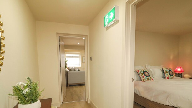 A hotel room with a bed, a bedside lamp, a small table with a plant, and a hallway leading to another room. - Suite 33A (Londonderry)
