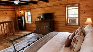 A wooden cabin bedroom with a bed, a television, a desk, and a ceiling fan.