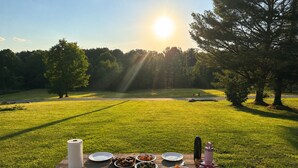 A picnic table with plates, food, and a bottle in a grassy field with trees and a sun in the background.