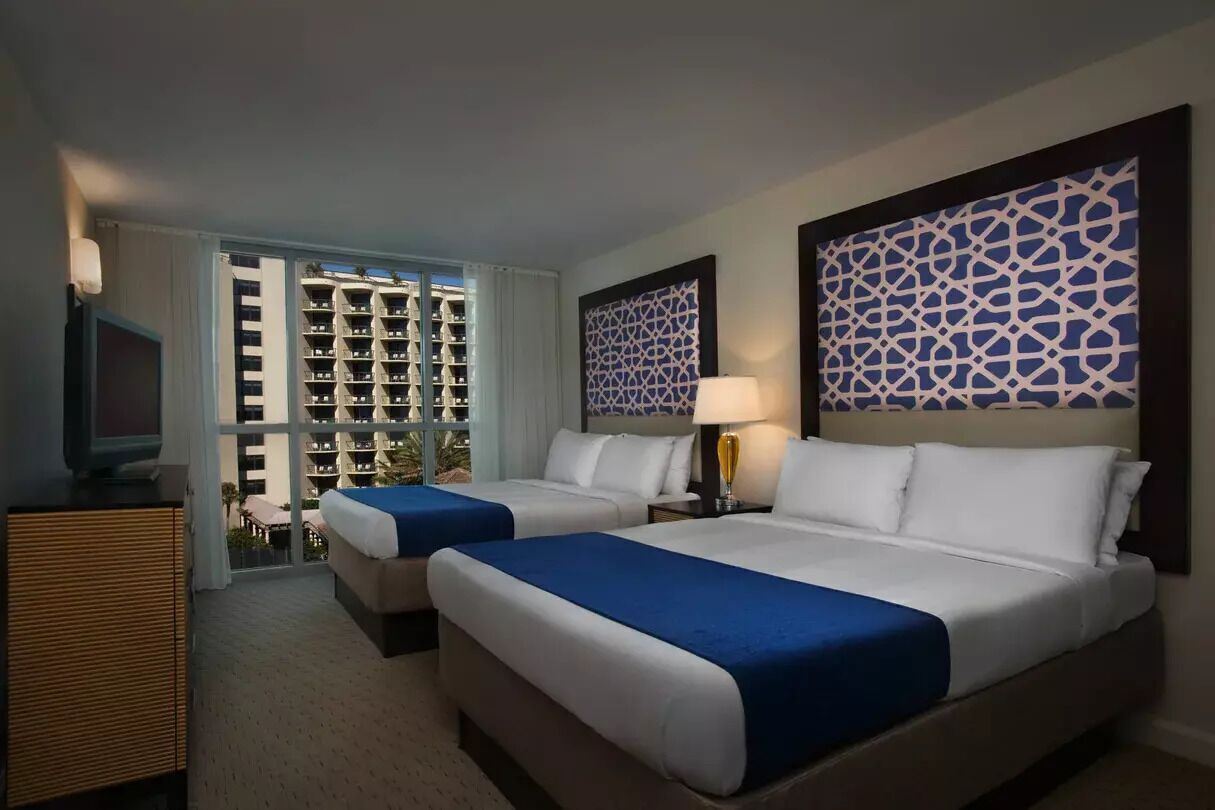 Habitación de hotel con dos camas, televisión, escritorio y vistas a un edificio con balcones.