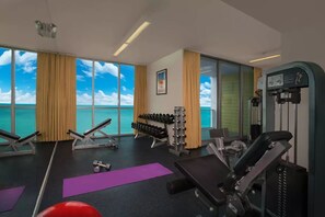 Ein Fitnessraum mit Trainingsgeräten, einer Hantelbank und großen Fenstern mit Meerblick.