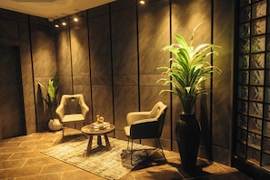 Quarto com decoração moderna, apresentando um sofá, uma cadeira, uma mesinha e uma grande planta em vaso.