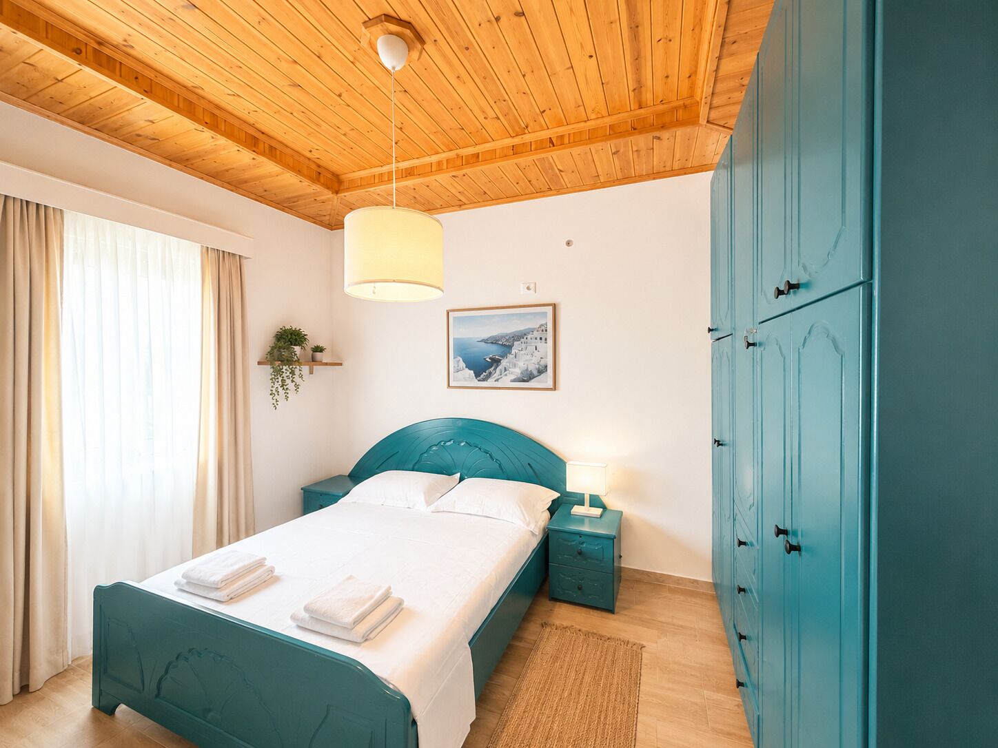 A bedroom with a wooden ceiling, a bed with a teal headboard, a nightstand with a lamp, a wardrobe, and a window with curtains.