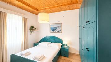 A bedroom with a wooden ceiling, a bed with a teal headboard, a nightstand with a lamp, a wardrobe, and a window with curtains.