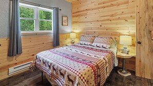 Una habitación con paneles de madera, una cama, dos lámparas de noche, una ventana con cortinas y un cuadro enmarcado en la pared.