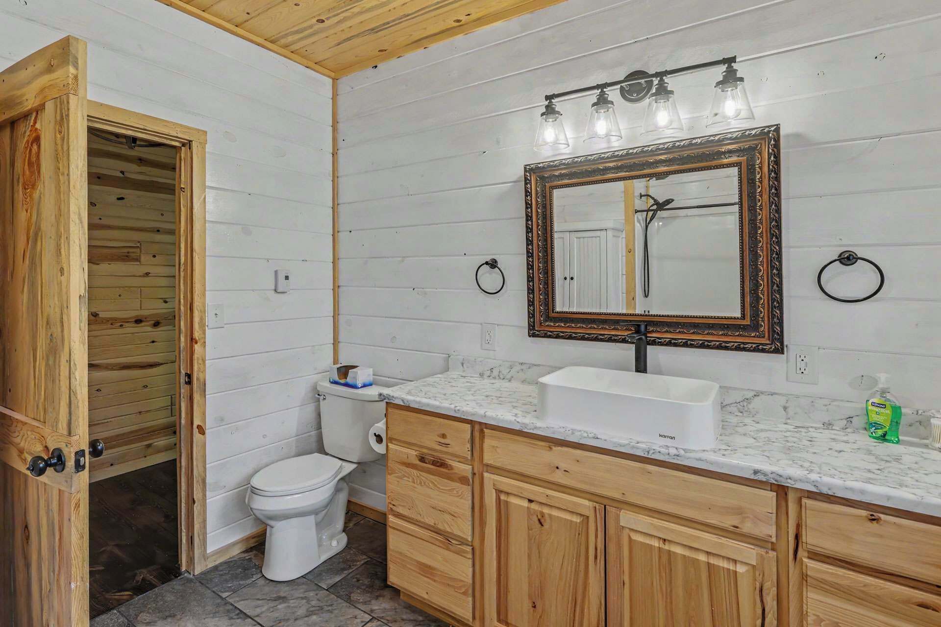 A bathroom with a wooden vanity, a white toilet, a marble countertop, and a mirror with a decorative frame.